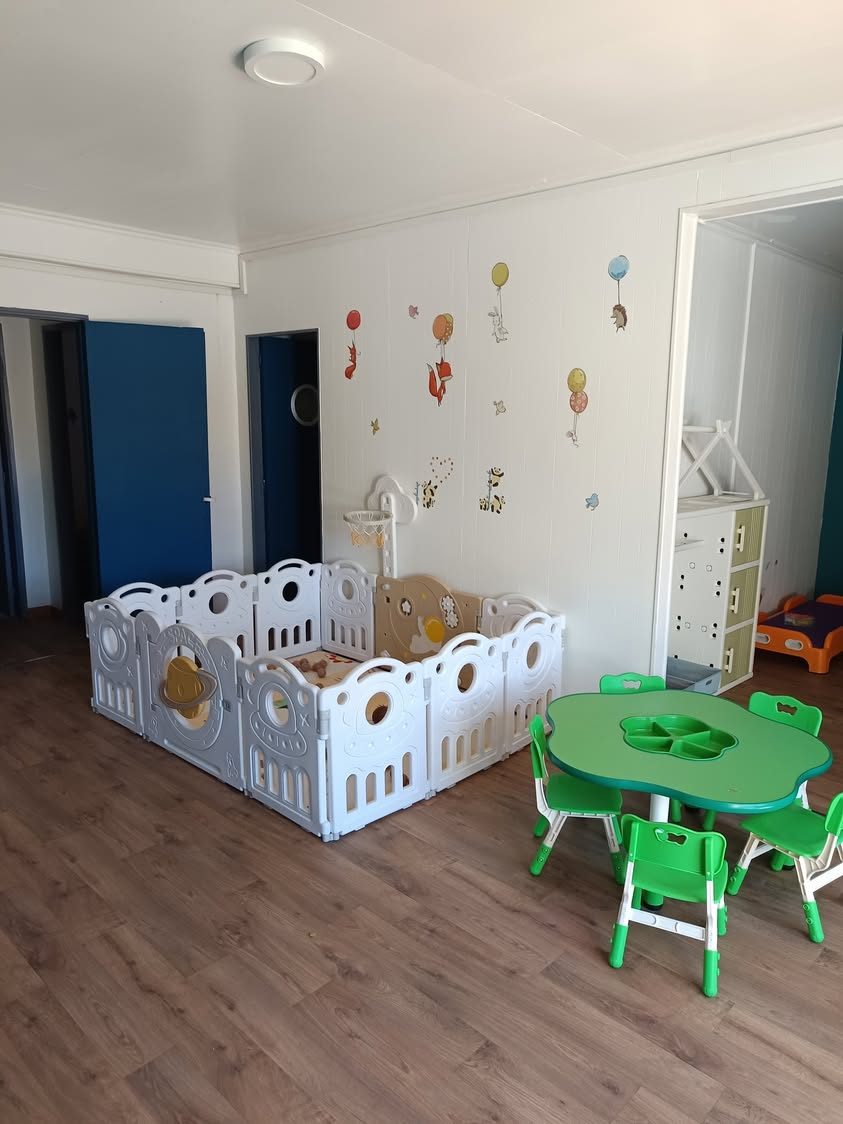 Salle de vie de la micro-crèche : parc de jeu blanc, table et chaises vertes, stickers au mur